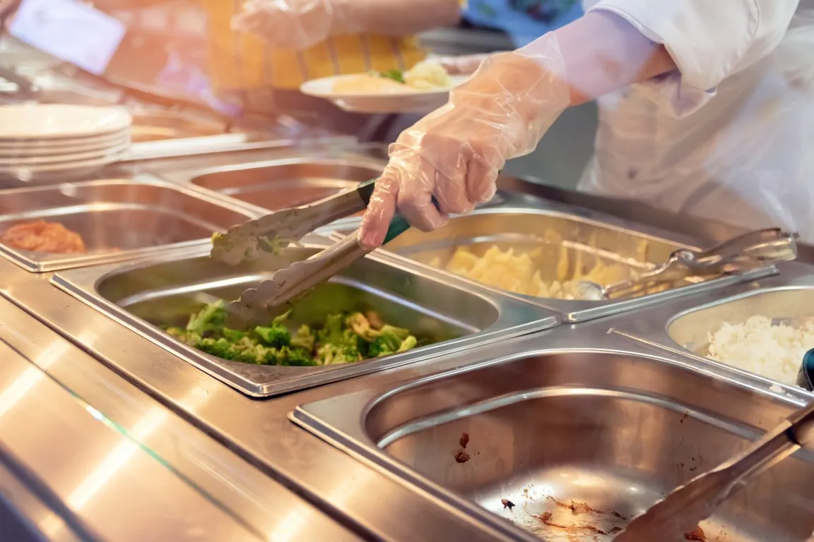 Mitarbeiterin mit Handschuhen portioniert Speisen an einer Essensausgabe